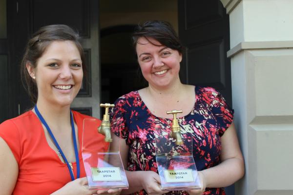 Two women dressed in red and blue holding awards that are made out of kitchen taps.