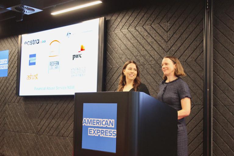 Two women stand smiling on at a podium wiht a screen behind them. On the podium is a large blue and white American Express logo