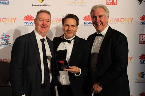 Three men in suits stand in a row hold a medal in a black box