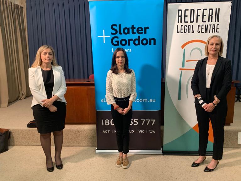 Three woman stand side by side each are holding their hands clasped in front of them. Behind them are banners that say Redfern Legal Centre and Slater and Gordon.