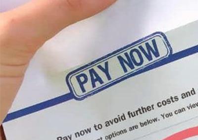 A closeup of a thumb holding a fines notice that says is large blue capital letters: "Pay Now"