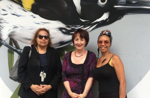 Three women stand in front of a mural of a painted bird. 