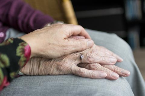 Two elderly women's hands. One hand wearing a diamond wedding ring rests on the arm of a grey couch with the other hand placed on in a gesture of support. 