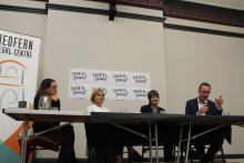 Three women and a man sit at a table up on a stage delivering a presentation in from of signs that say "Safe and Sound"