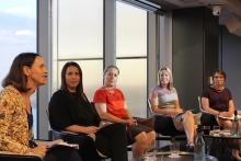 Group of four women seated on a stage in front of a window prenting a discussion to a crowd. The woman in the foreground of the photo woman wears a headset microphone.