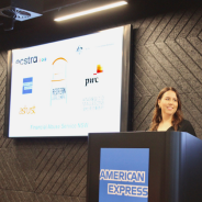 A woman with long brown hair stands at a pulpit with a blue Amex logo on it in front of a screen displaying a group of logos
