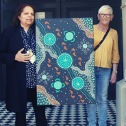 Two women stand either side of a black green and orange Aboriginal painting
