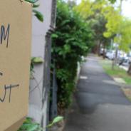 Text reads "Room for Rent" on piece of cardboard attached to fence on suburban street